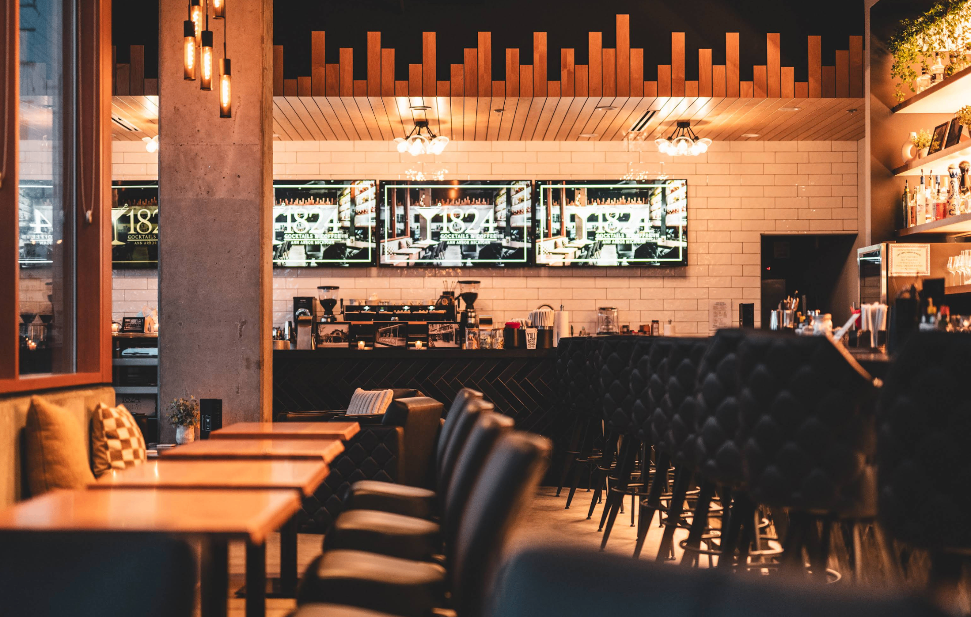 Warm interior view of 1824 Cocktails & Coffee with leather seating, glowing bar, and illuminated screens